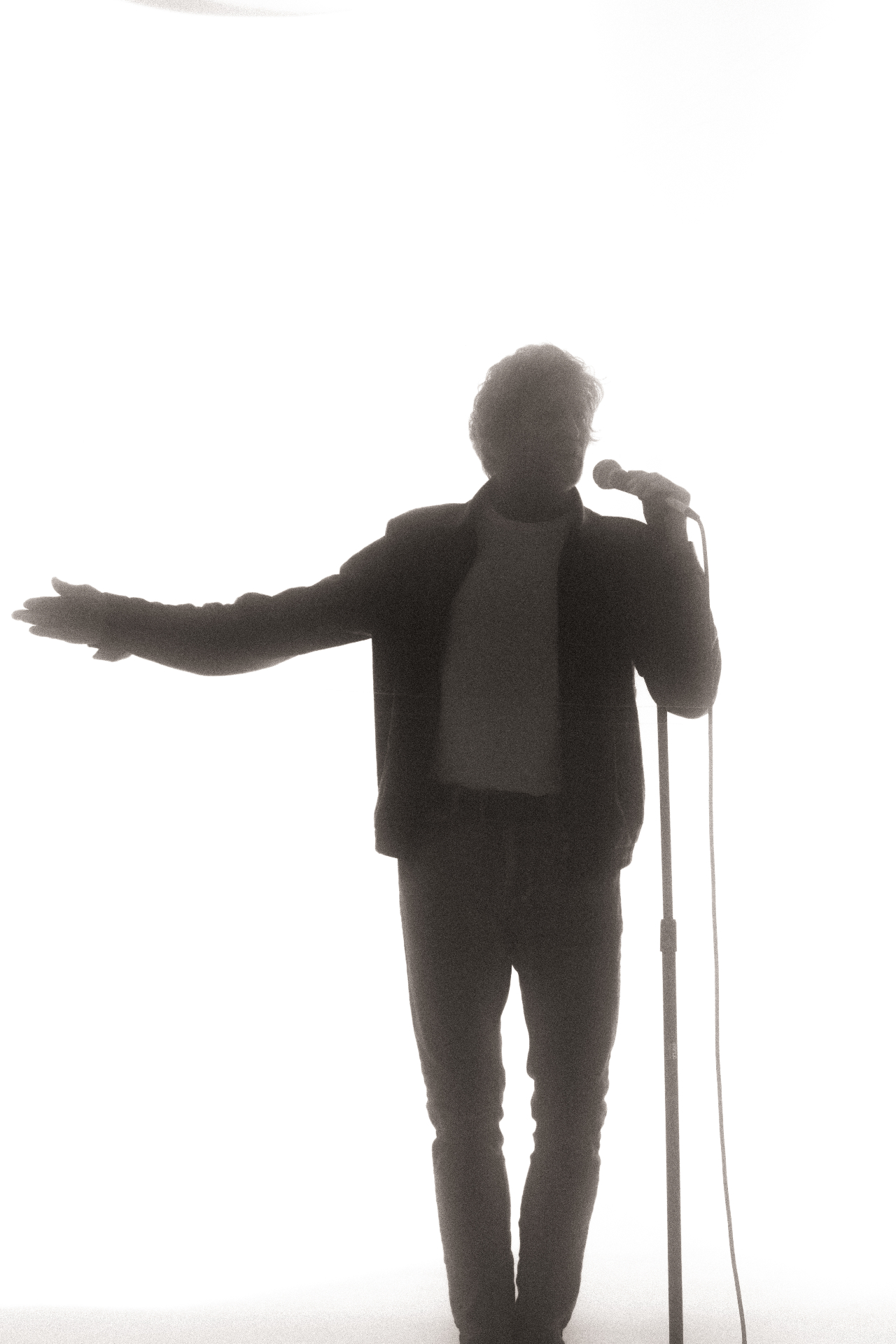 Gregg Hodge standing at a microphone under a single spotlight